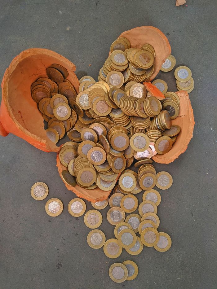 A shattered clay piggy bank with ten rupee coins scattered on the floor.