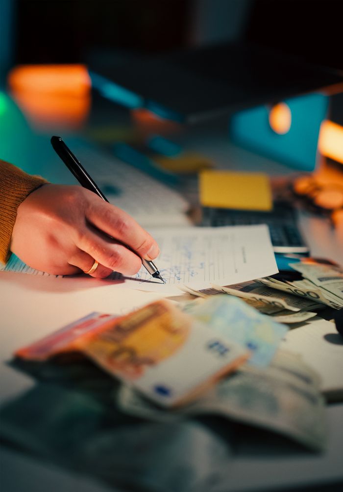 Close-up of a hand writing on documents with euro notes scattered on a desk, focusing on finance and business.